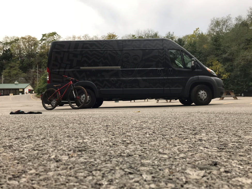 Right side view of a red bike leaning on a rear wheel well of a black Surly van, at a parking with trees behind