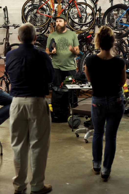 The backsides of 2 people listening to a person to a person facing them with bikes hanging from racks in the background