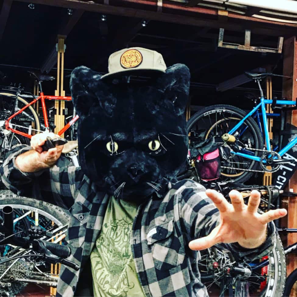 Front view of person in a karate pose, wearing a large black panther head, in front of a garage with bikes around