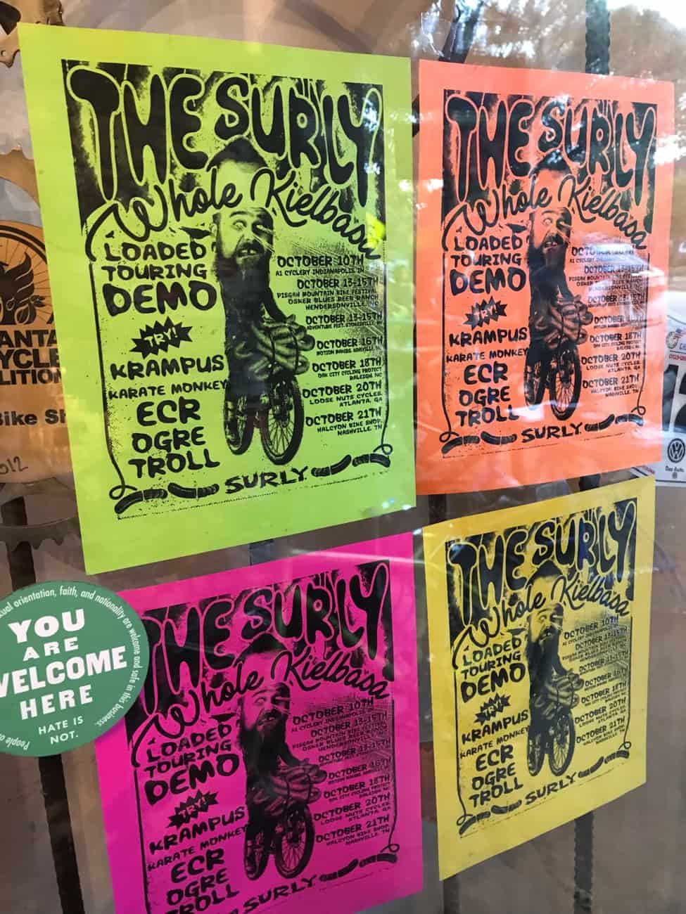 Four colorful 'The Surly Whole Kielbasa', flyers taped to a store front window 