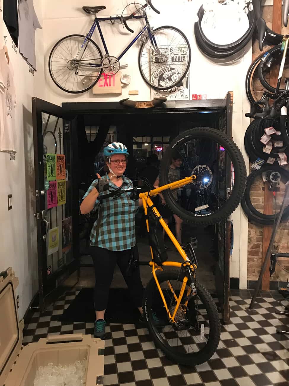 Front view of a cyclist giving a thumbs up at a bike shop, while holding a bike with front wheel popped off the ground
