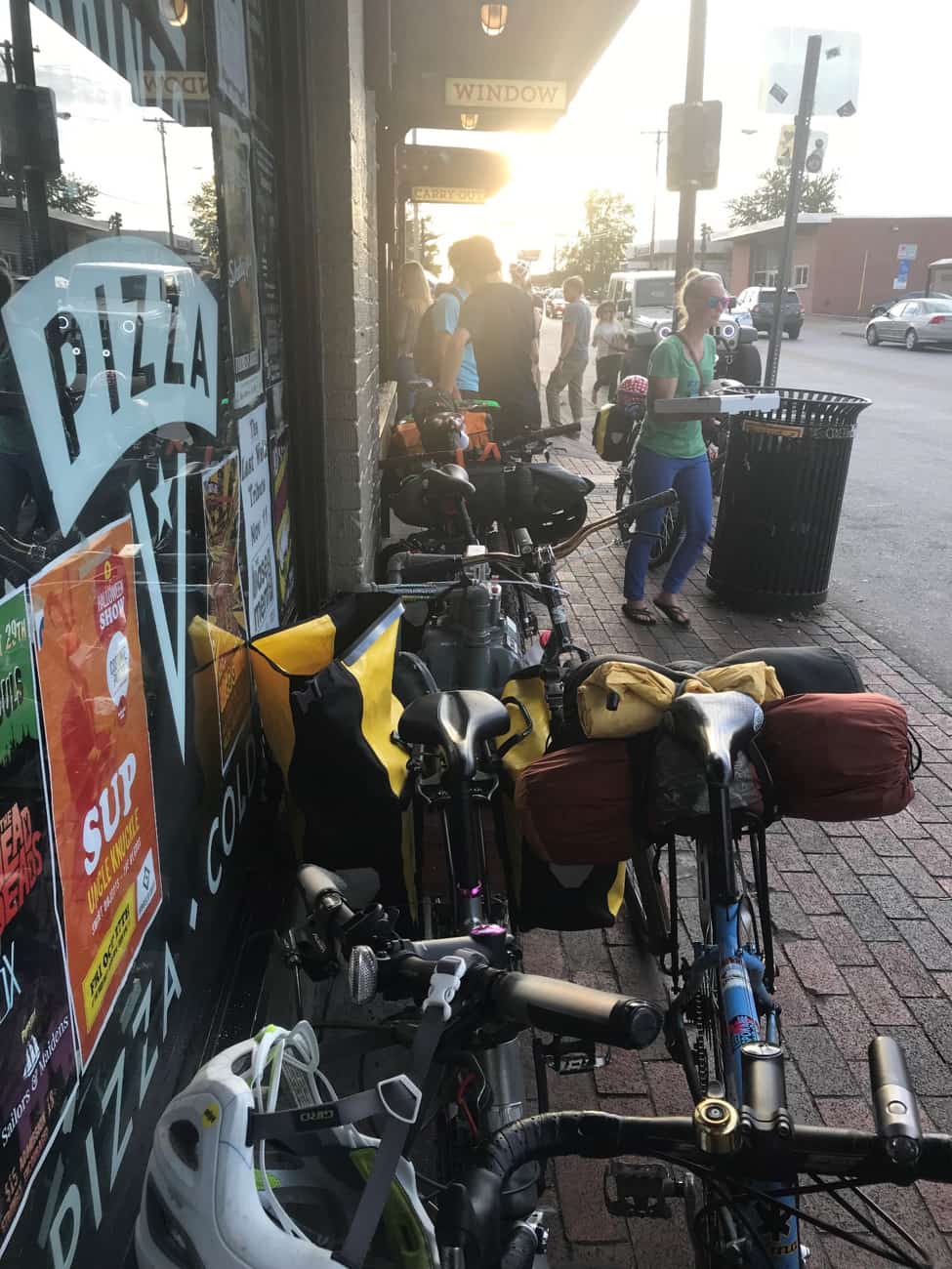 Bikes lined up and parked on a red brick sidewalk, next to the window of a pizza shop