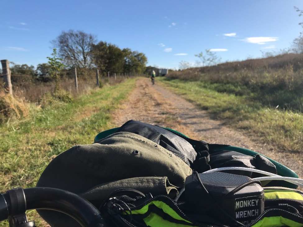 View from the top of a bike handlebar gear pack, facing down a country gravel road  with thick grass to the sides of it
