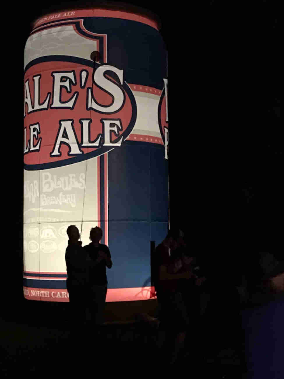 Silhouettes of people standing in front of a large, lighted inflatable beer can at night 