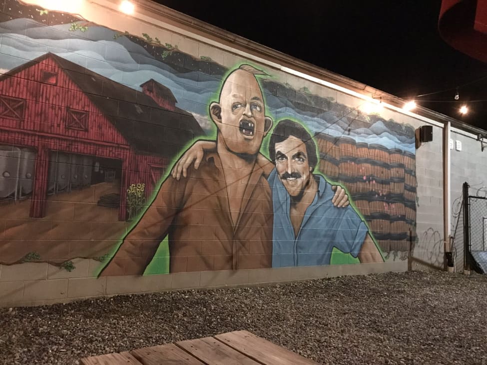 Nighttime view of a mural on a wall showing Lar, from Goonies, and Tom Selleck with their arms on each others shoulders