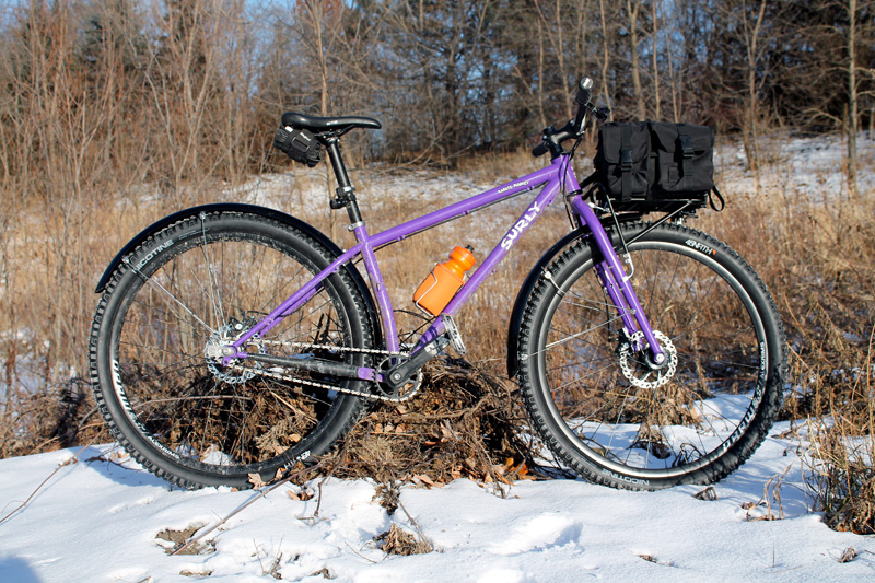 Right profile of a Surly Karate Monkey bike with fenders, 8 Pack rack with gear pack on top, on a snowy field with brush