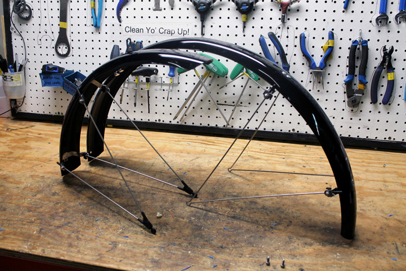 Side view of a black front and back fenders, with mounts, on top of a workbench with tool loaded pegboard behind