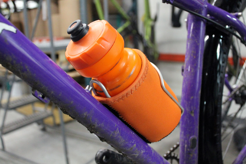Close up view of a orange bottle inside a bottle cage, mounted to the down tube of a Surly Karate Monkey bike, purple