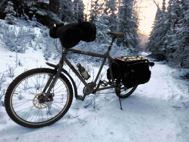 Left side view of an olive colored Surly Big Dummy bike with a front pack, parked on a snow covered trail in the forest