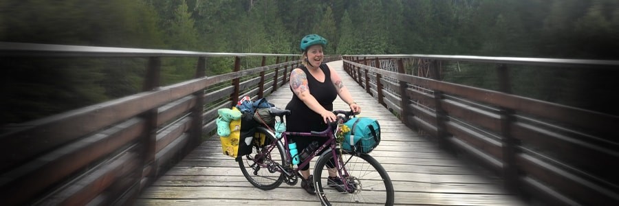 Cyclist stands on left side of fully geared Surly Straggler bike on wood bridge trail with pine trees in background