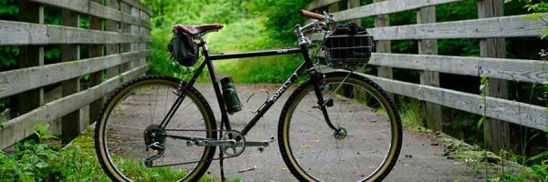 森の中の舗装された橋の上に停めた前カゴを付けた Surly Pack Rat 自転車の側面