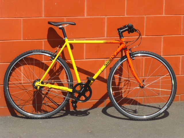 Right side view of a red and yellow Surly Cross Check bike, parked on a sidewalk, against a red block wall