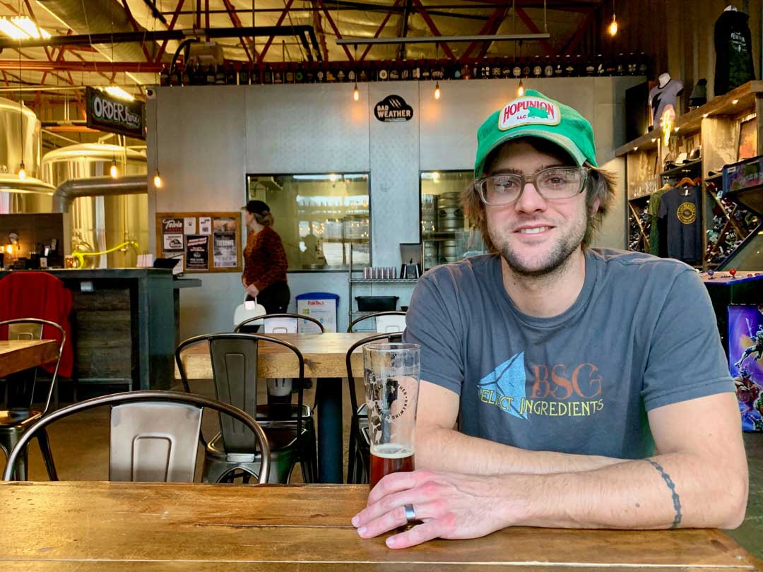 Head Brewer, Andy, wearing a green baseball cap and glasses sits at a table with a glass of beer at a brewery