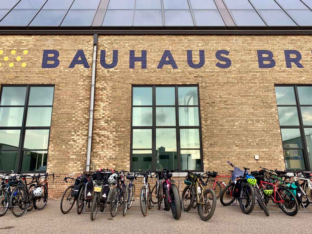 Cluster of bikes loaded with gear in front of three large windows on a brick Bauhaus Brewery building with glass roof