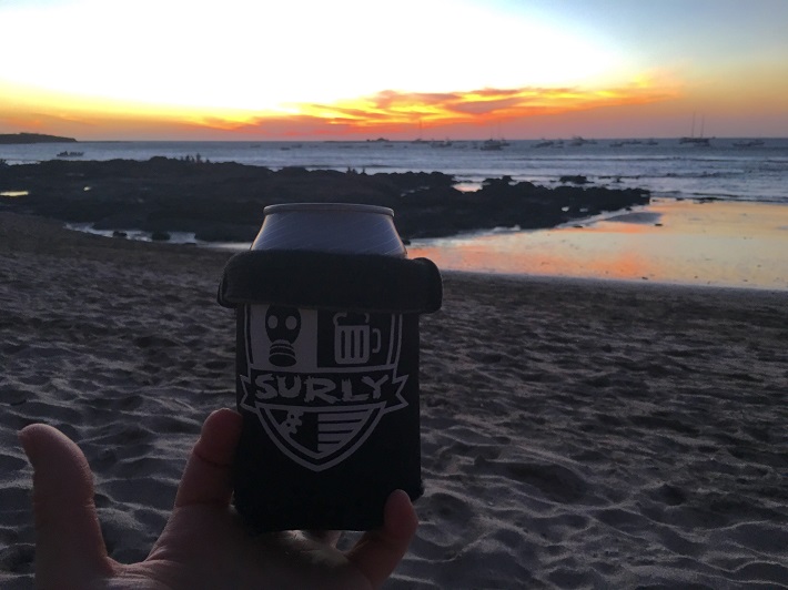 A hand holding up a Surly can cooler with a beach, the ocean and golden clouds above at dusk