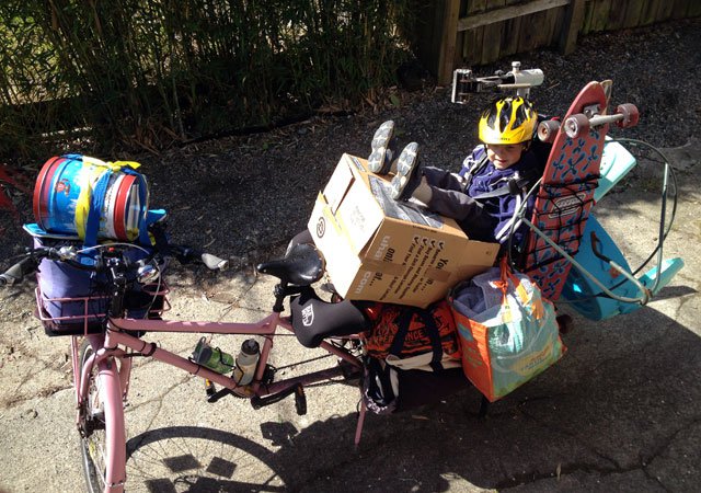 Downward, left side view of a pink Surly Big Dummy bike, loaded up, and a child sitting on a cardboard box, in the back
