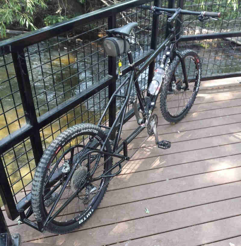 Rear, right side view of an olive drab Big Dummy bike, leaning against a handrail, on a wood deck over a stream