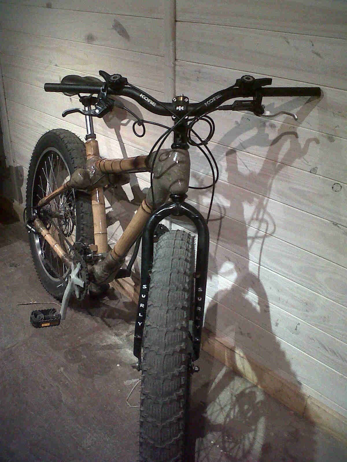 Front, right side view of a bike with a bamboo frame, leaning against a white painted wood wall, inside of a room