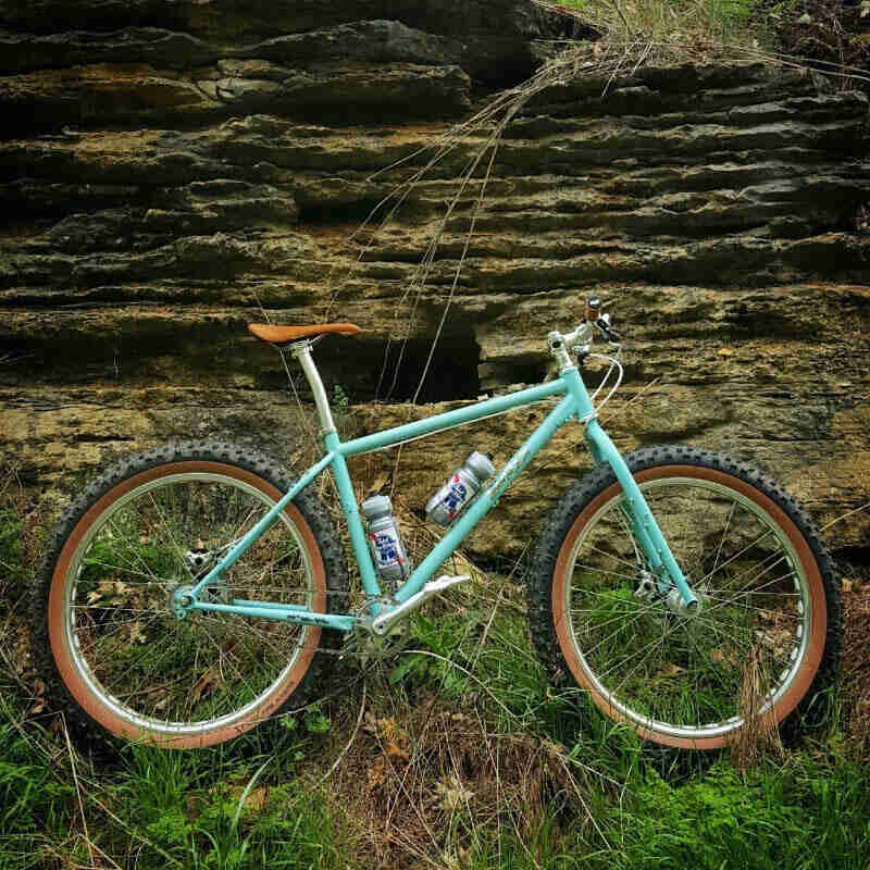 Right profile of a turquoise Surly fat bike with PBR water bottles, standing in weeds, in front of a rock ledge wall