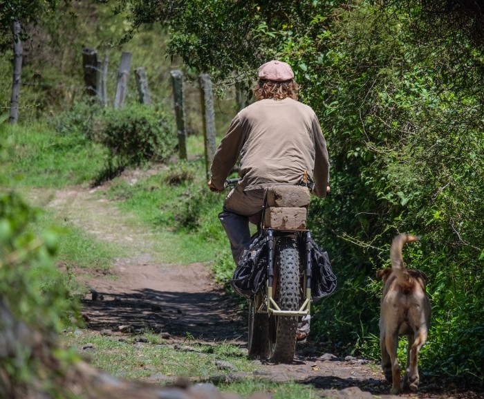 Rear view of a cyclist riding down a dirt trail alongside a fence, on a Surly Big Fat Dummy bike, with dog following