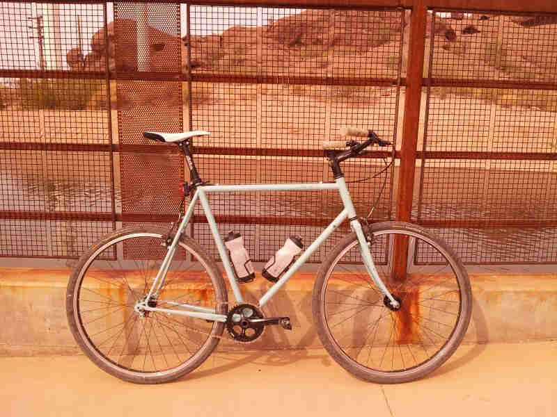 Right side view of a mint Surly bike, leaning against a steel grid fence, on a sidewalk, with water and hills behind it