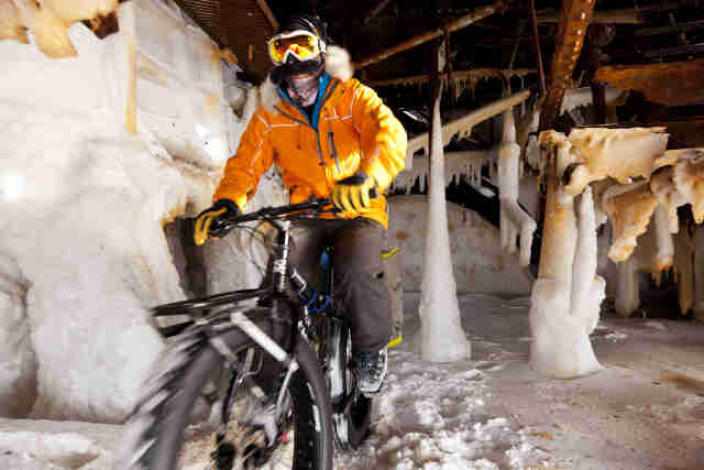 Front, left side view of a cyclist in winter outerwear, riding a Surly Moonlander fat bike in a steel structure on ice