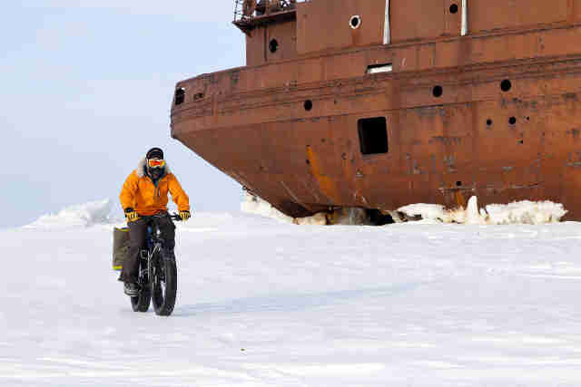 Front view of a cyclist in winter attire, riding a Surly Moonlander fat bike on a frozen field with grounded ship behind