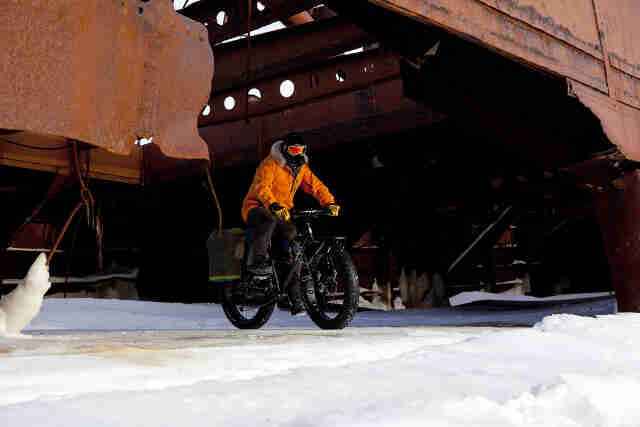 Front, right side view of a cyclist, wearing winter attire, riding a Surly Moonlander fat bike out of a structure on ice