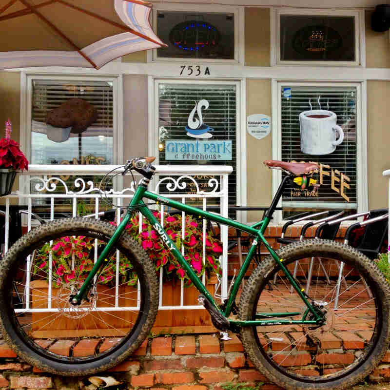 Left side view of a Surly Krampus bike, green, on a red brick sidewalk in front of a coffee shop