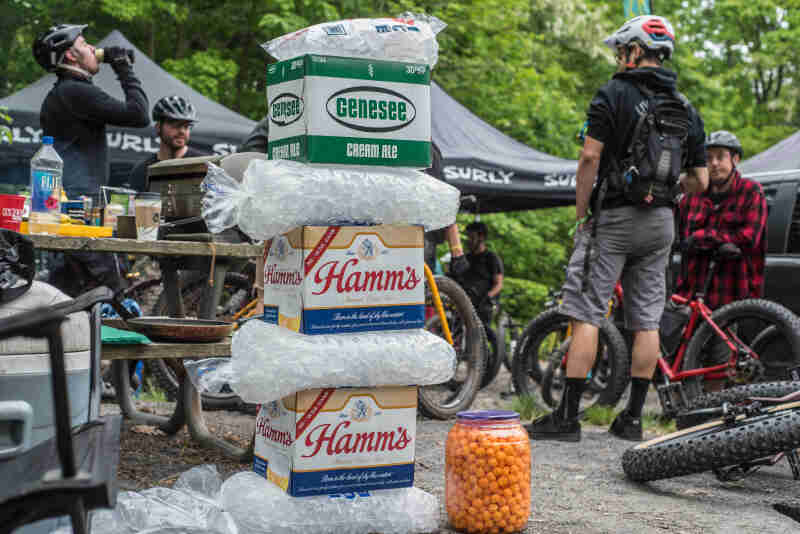 A stack of ice and cases of beer, with cyclists standing to the sides, and canopies and trees in the background