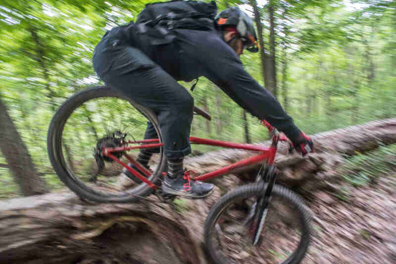 Right side view of a cyclist, on a red Surly bike, riding over a downed tree, in the woods