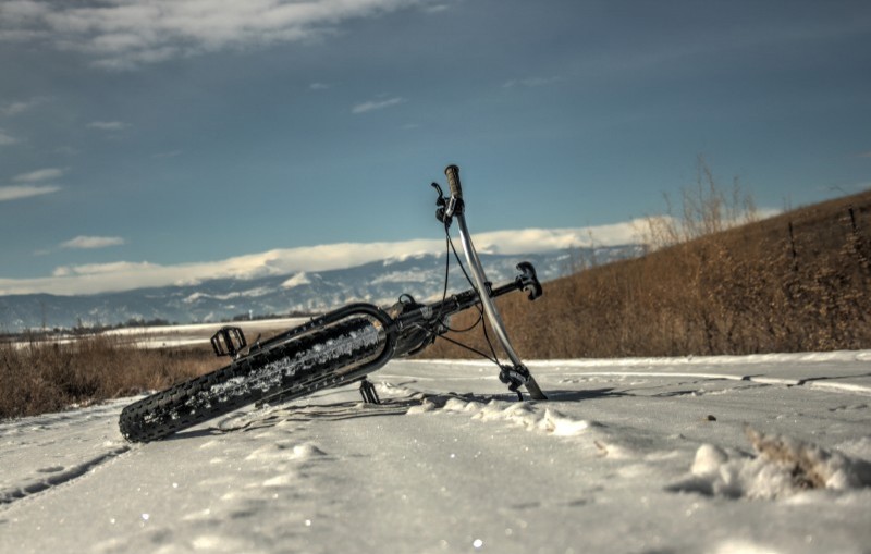 Front, ground level view of a black Surly fat bike, laying on left side, on a snowy road with mountains in background