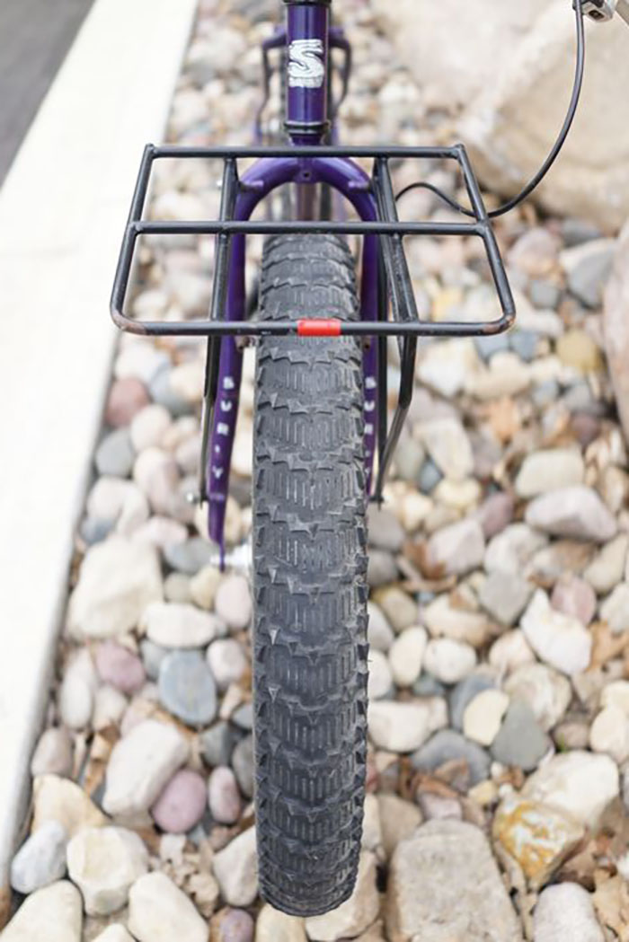 Partial front view of a purple Surly Pugsley fat bike head tube, forks, carry rack and fat tire on stones