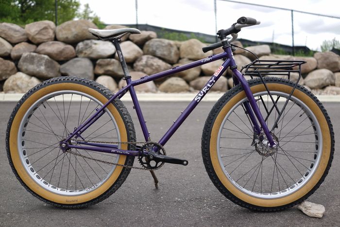 Left side view of a purple Surly Pugsley fat bike on pavement, with a boulder wall and chain link fence above it
