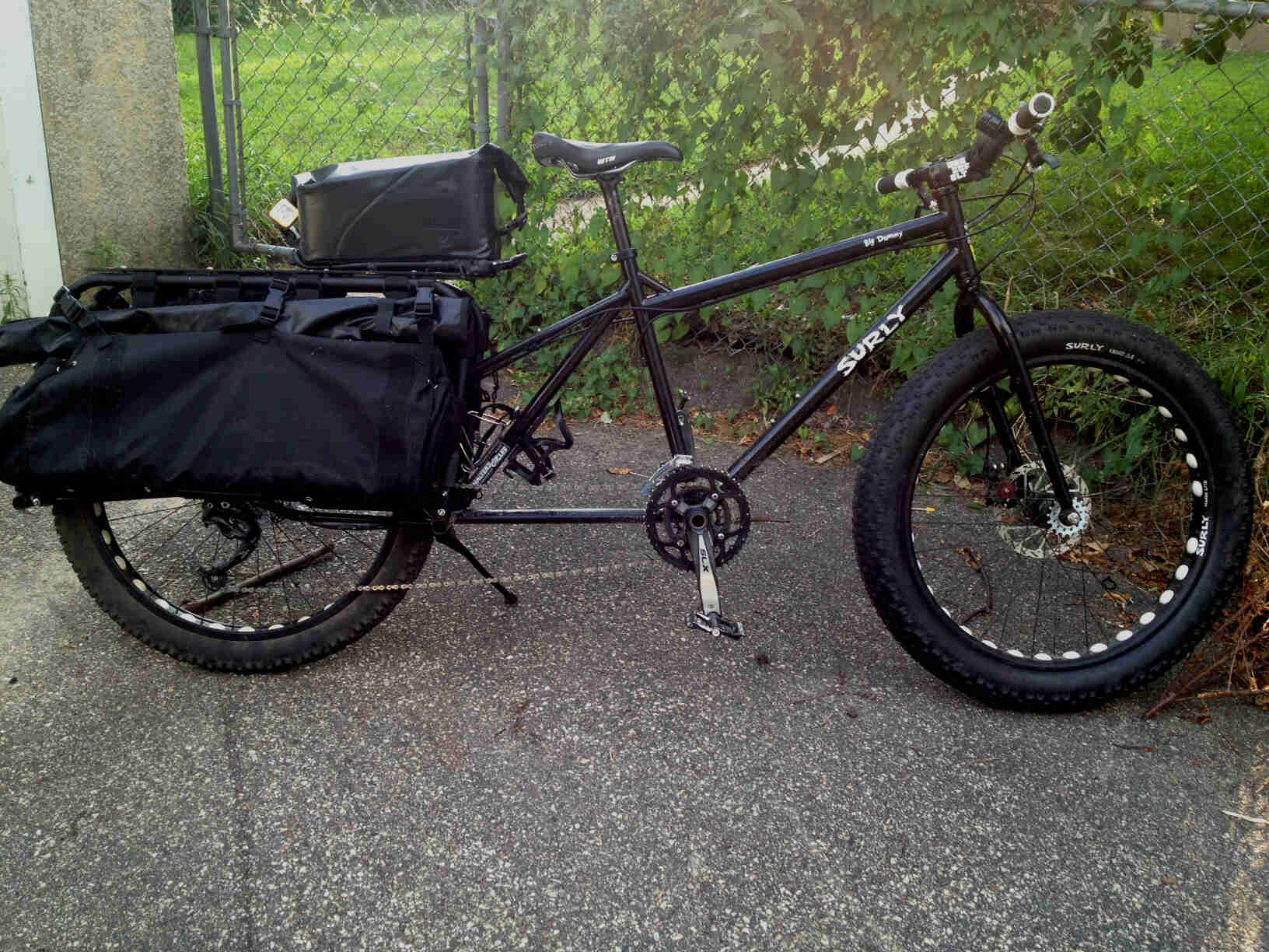 Right side view of a black Surly Big Dummy bike with a fat front wheel, on pavement, with a chain link fence behind