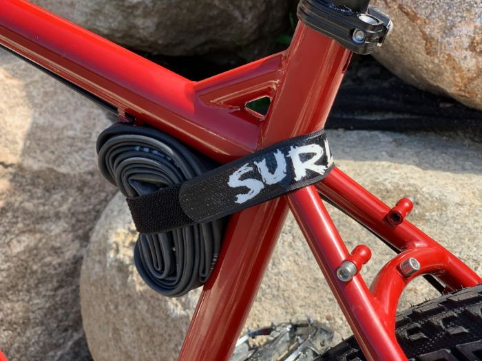 Rolled up bike inner tube attached to a red bike frame with a Surly Whiplash frame strap with boulders behind