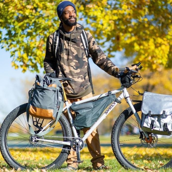 A cyclist wearing a camouflage sweatshirt stares off while holding a white Surly bike with gear attached