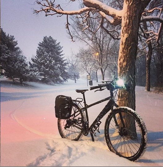 A black Surly ECR bike with headlight on sits against a tree at night in a snow covered field with pine trees behind it