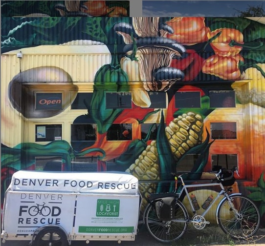 A bike with and a Denver Food Rescue trailer behind it is parked aside steel building painted in colorful vegetables