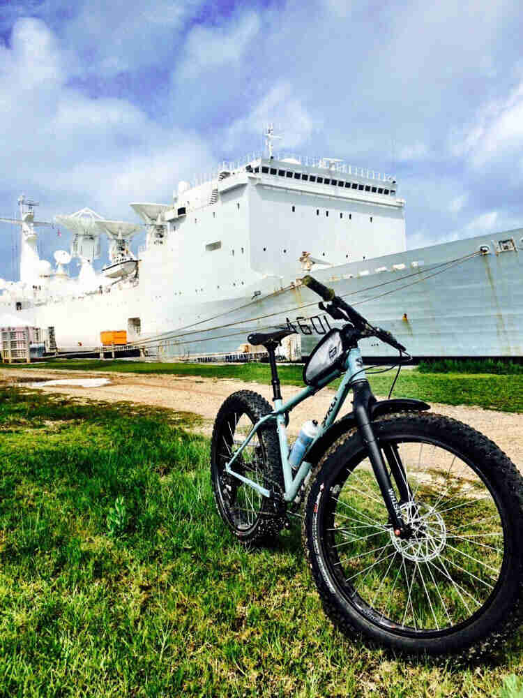 Front left view of a Surly Wednesday MY fat bike parked in grass, next to a gravel road, with a ship in the background