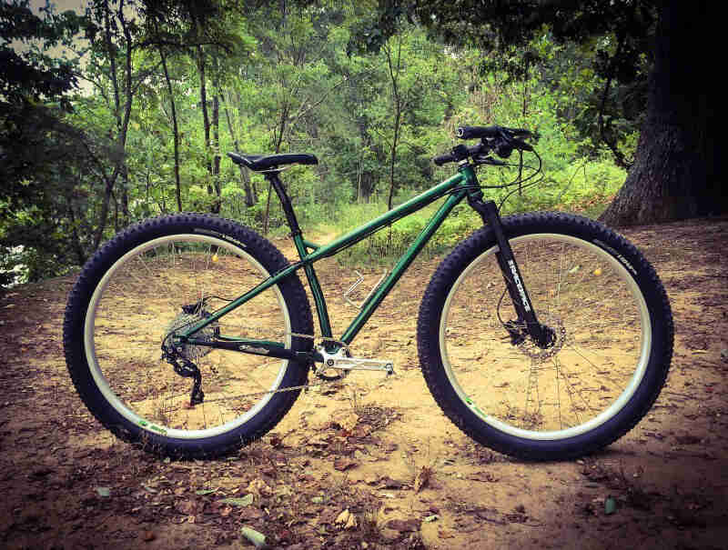 Right side view of a Surly Krampus bike, green, parked on a dirt lot, with a thick forest in the background
