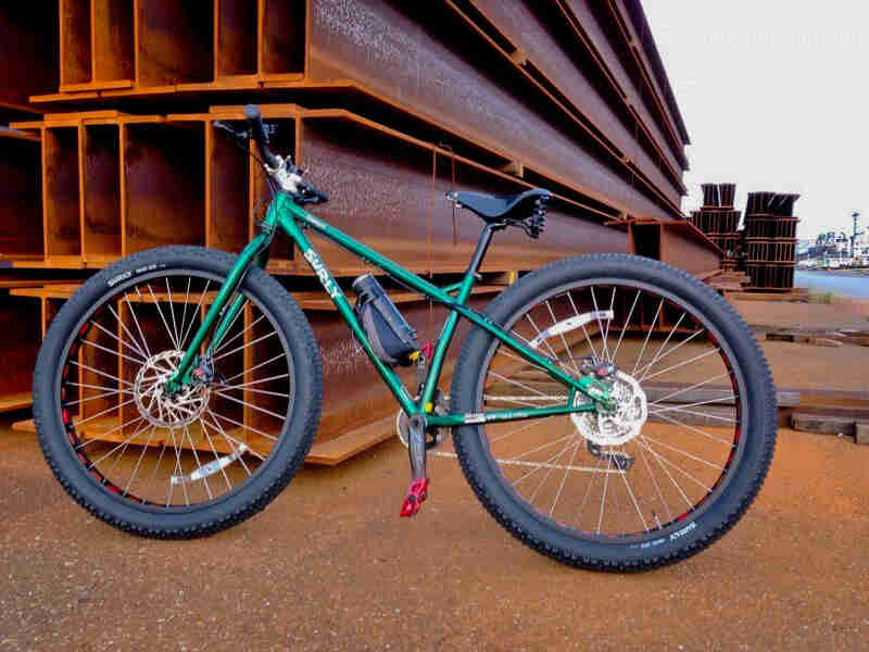 Left side view of a Surly Krampus bike, green, leaning on a stack of beams at a steel yard