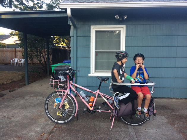 Left side view of a Surly Big Dummy bike, pink, with two children sitting in the rear rack  in front of a house
