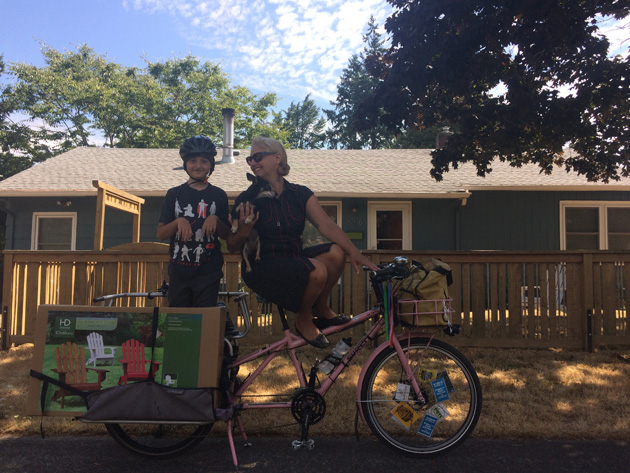 Person seat on a Surly Big Dummy bike, pink, with a child standing up in the rear rack  and house shown in background
