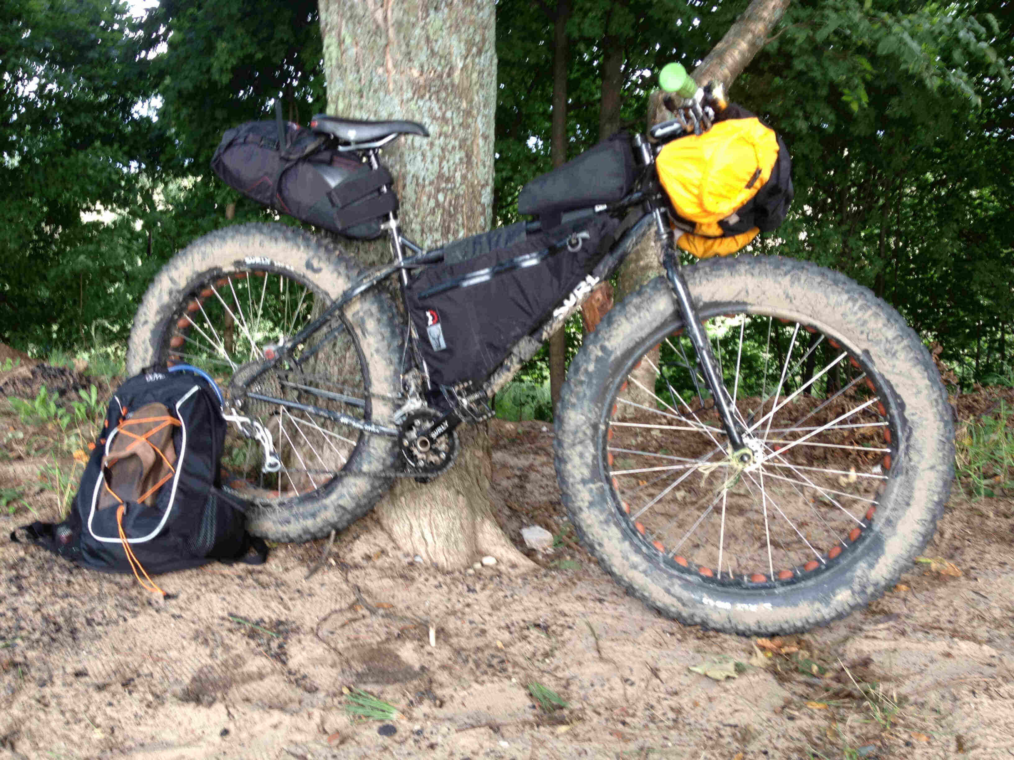 Right side view of a Surly Moonlander fat bike with gear, leaning against a tree on dirt ground, with trees behind it