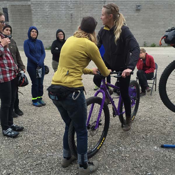 Backside view of a person holding the handlebar of a purple Surly bike with rider, in a gravel lot, with people watching