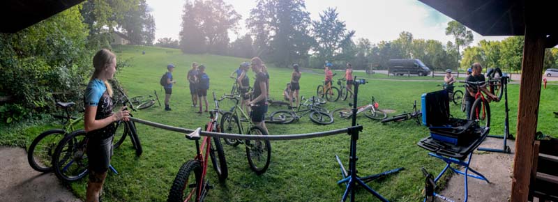 Panoramic view of a group of cyclists standing in a grassy field, with their bikes scattered around, near a park gazebo