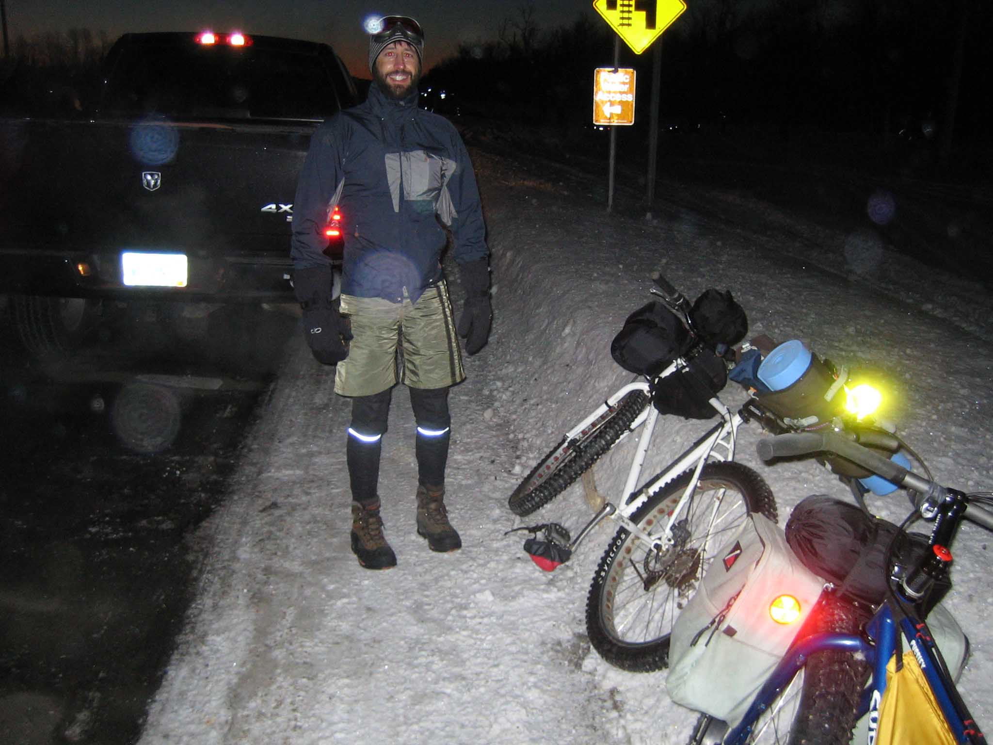 A smiling cyclist standing on a snowy roadside with 2 bikes laying down, at night