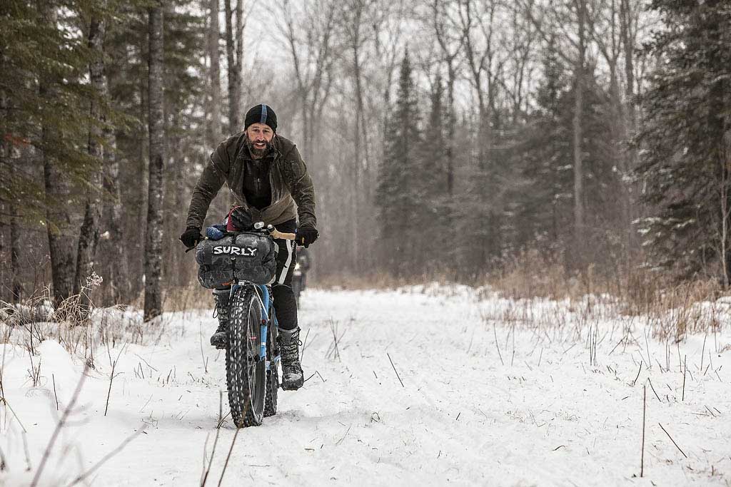 Front view of a cyclist riding a fat bike with a Surly front pack, on a snow covered trail in the woods