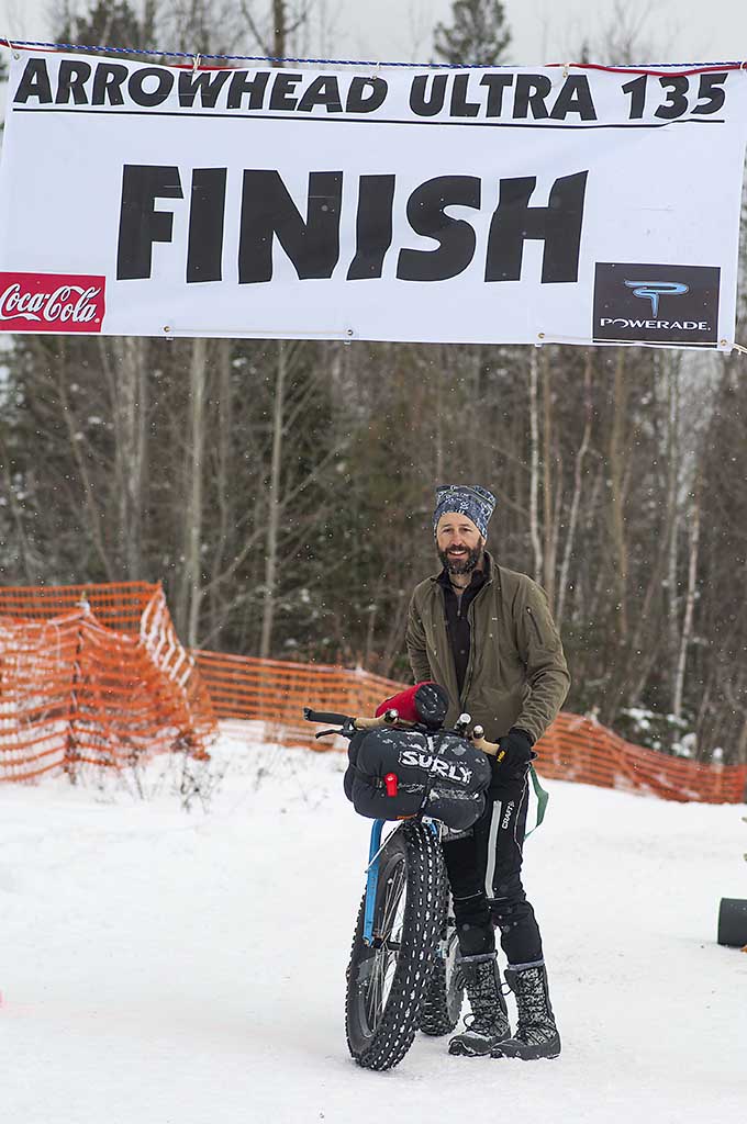 Front view of a cyclist standing next to a blue fat bike with a Surly front pack, at a snowy finish line